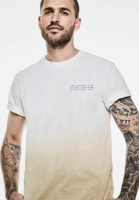 Vit t-shirt med en gradient till ljusbeige, korta ärmar och en liten svart textdesign på bröstet. Mjuk bomullsmaterial.