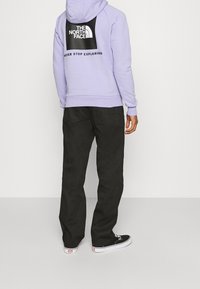 Persoon in een lila hoodie met het 'The North Face' logo en de slogan 'Never Stop Exploring', zwarte broek en zwarte Vans sneakers.