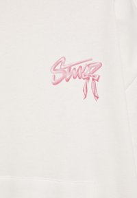 Tissu blanc avec texte stylisé rose et un motif de nœud en haut à gauche, au-dessus d'une ligne de couture horizontale.