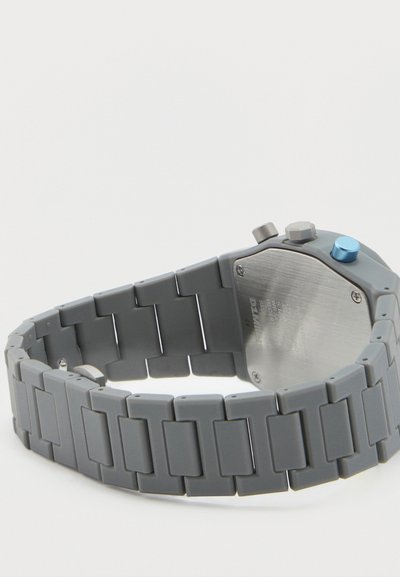 Γκρι μπρασελέ ρολογιού με συνδέσμους, με ανοξείδωτο ατσάλι στην πίσω πλευρά και τρία κουμπιά, εκ των οποίων ένα μπλε, παρουσιάζεται σε λευκό φόντο.