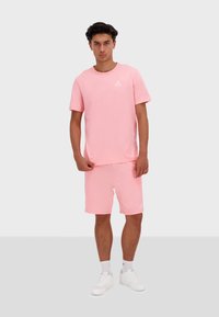 Completo in cotone di colore rosa chiaro costituito da una T-shirt e da pantaloni corti, con scollatura rotonda, maniche corte e logo bianco sul petto, abbinato a sneakers bianche.