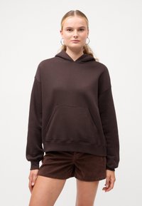 Hanorac maro dintr-un material moale, cu un buzunar frontal, umeri căzuți și glugă cu șnur, asociat cu pantaloni scurți maro.