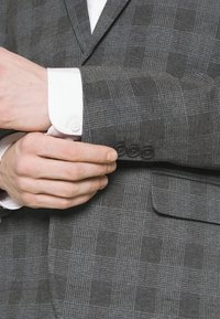 Grijze ruitjes blazer van een gestructureerde stof, met drie zwarte knopen op de manchetten en een wit overhemd eronder.