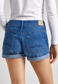 Jeansshorts med uppvikta fållar, medelhav blå färg, med två bakfickor med texturerad söm och en brunt läderlogotyp.