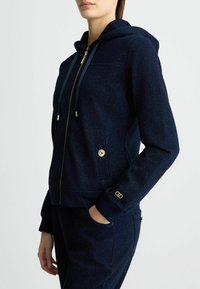 Felpa con zip in navy realizzata in tessuto testurizzato, presenta una zip dorata, cordini, tasche laterali con bottoni dorati e polsini a costine.
