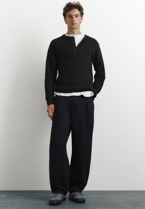 Un giovane uomo è in piedi contro un muro bianco, indossa una henley nera a maniche lunghe sopra una camicia bianca, pantaloni larghi scuri e sneaker grigie.