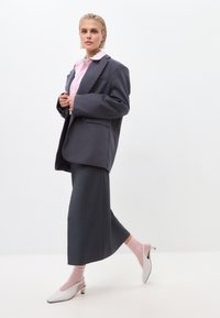 Blazer gris surdimensionné sur une chemise rose pâle, assorti à une jupe grise et des chaussettes rose pâle. Des escarpins blancs à lanières complètent le look.