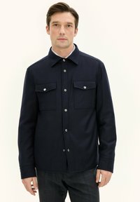 Giacca camicia blu navy con colletto a punta, due tasche sul petto e bottoni a pressione argentati. Il tessuto sembra liscio con un taglio strutturato.