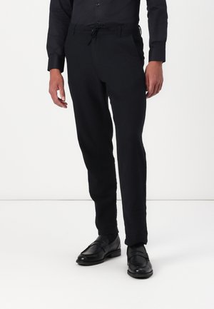 Mężczyzna ubrany w czarne eleganckie spodnie, czarną elegancką koszulę oraz czarne skórzane mokasyny, stojący na białym tle.