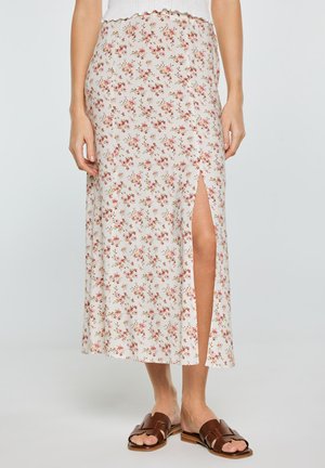 Kobieta nosząca białą kwiatową spódnicę midi z przednim rozcięciem, w połączeniu z brązowymi płaskimi sandałami, stojąca na tle jednolitego jasnego tła.