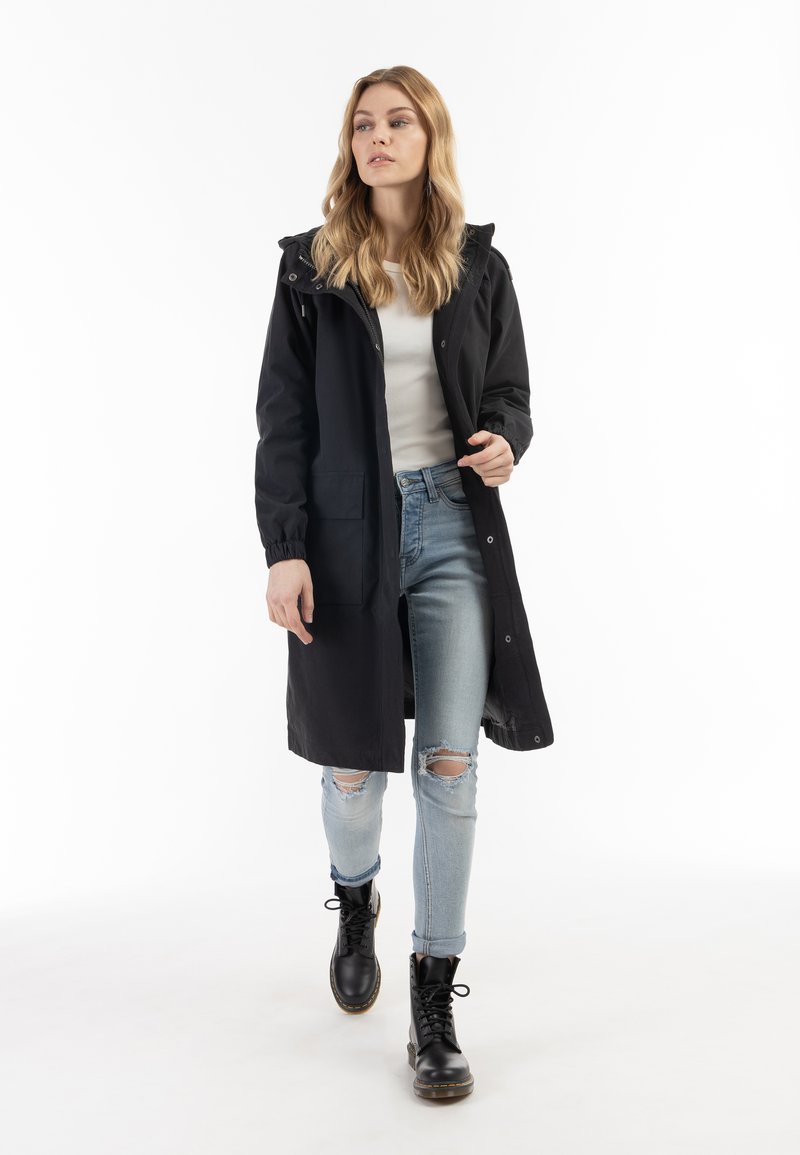Cappotto lungo impermeabile nero con cappuccio, chiusura frontale a pressione e tasche laterali, indossato sopra una maglia bianca e jeans blu strappati, abbinato a stivali neri.