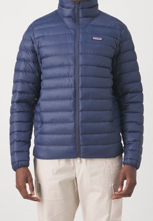 Homme portant une veste matelassée Patagonia bleu marine zippée et un pantalon beige clair, mains détendues le long du corps, fond uni.