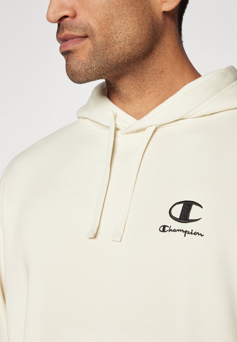 Sudadera color crema hecha de mezcla de algodón, con un bolsillo canguro, cordones y un logo de Champion bordado en negro en el pecho.