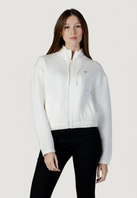 Sudadera blanca corta con cremallera y cuello alto, con un detalle de logo en el pecho y puños elásticos. Combinada con pantalones negros.