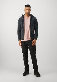 Fleece bunda tmavosivej farby s textúrovaným vzorom, kapucňou, zipsovými vreckami, skombinovaná so svetloružovou košeľou a čiernymi nohavicami typu cargo. Celkom to dopĺňajú čierne topánky.