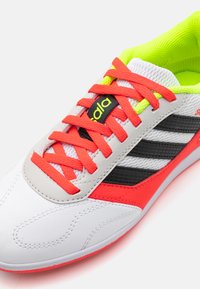 Basket blanc avec un éclairage néon vert vif, des lacets rouges et des rayures noires. Présente un matériau texturé et un design à bout rond.