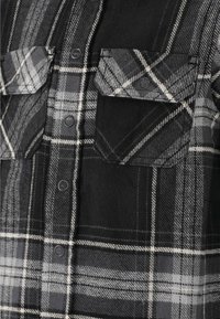 Chemise en flanelle à carreaux noirs et gris avec un devant à boutons, deux poches poitrine et des détails en motif à carreaux contrastants. Texture douce, design décontracté.