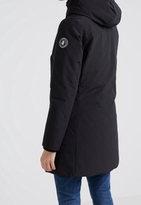 Sort isoleret parka med hætte, udstyret med et cirkulært logo-patch på den øverste arm og ribbede ærmer. Glat ydre overflade, mellem-længde design.
