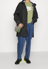 Svart vattentät jacka med huva, grön T-shirt med vit logotyp, blå avslappnade jeans och svarta skor med neongröna sulor.