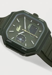 Τετράγωνο πράσινο ρολόι χειρός Lacoste με αναλογική και ψηφιακή ένδειξη, καντράν ημέρας της εβδομάδας και κουμπιά με ετικέτες στις άκρες της θήκης.