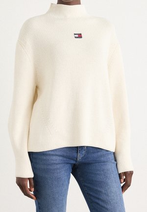 Person iført en kremfarvet ribstrikket sweater med høj hals og et Tommy Jeans-logo, kombineret med blå denimjeans.