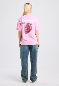 Pink cotton t-shirt featuring a large strawberry graphic and text "Taste the Season" on the back. Loose fit with short sleeves.