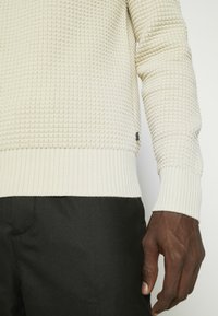 Hell gefärbter, strukturierter Pullover mit geripptem Saum und Bündchen, versehen mit einem kleinen Logotag an der Seite. Über dunklen Hosen getragen.