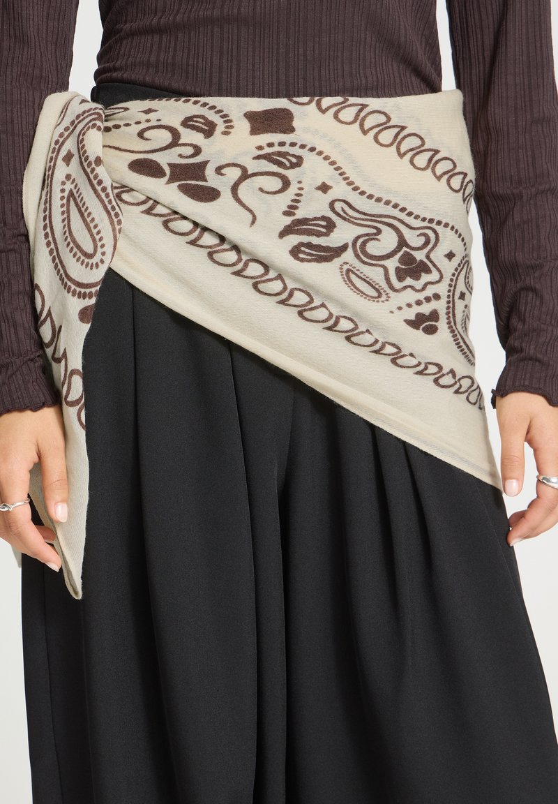 Personne portant une jupe plissée noire et un haut côtelé marron avec une écharpe crème et marron à motifs nouée autour de la taille.