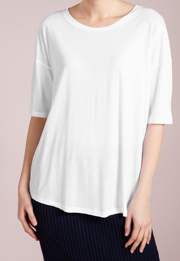 Femme portant une chemise blanche ample à manches courtes avec un col rond et une jupe côtelée foncée, debout devant un fond clair.