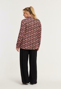 Langärmlige, gemusterte Jacke in Schwarz und Pink mit ovalen Designs, kombiniert mit schwarzen weiten Hosen und High Heels.