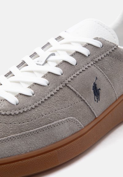Γκρι σουέτ αθλητικό παπούτσι με λευκά κορδόνια, καφέ λαστιχένια σόλα και σκούρο κεντημένο λογότυπο παίκτη πόλο στην πλαϊνή επιφάνεια.