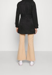 Persona de espaldas con un abrigo trench negro, pantalones acampanados beige y zapatillas Converse blancas en un fondo sencillo.