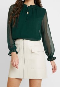 Blusa verde trasparente con maniche lunghe a volant e dettaglio colletto con laccetto, abbinata a una mini gonna in pelle color crema con zip frontale e tasca.