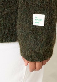 Tamsiai žalias megztinis su raštuotais rankogaliais, su balta etikete, kurioje žaliais užrašais parašyta "LA MAISON AMV." Textūrinis, storas audinys.