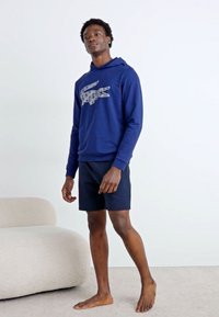 Marineblaues Hoodie mit einem Krokodil-Logo, kombiniert mit schwarzen Shorts. Der Stoff wirkt weich und leicht. Model steht barfuß.