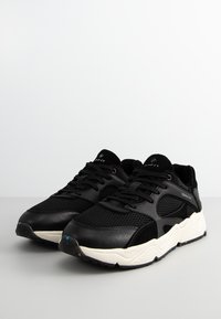 Ein Paar schwarze GANT-Sneaker mit weißen, klobigen Sohlen, Obermaterial aus Mesh und Leder sowie schwarzen Schnürsenkeln auf einer hellgrauen Oberfläche.