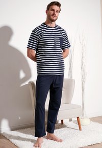 Marineblauw en witte gestreepte shirt met korte mouwen en een borstzak, gecombineerd met effen marineblauwe broek. Model staat blootvoets op een zachte witte tapijt.