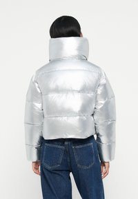 Silver puffer jacket with high collar, horizontal stitching, and a shiny, reflective texture. Worn with dark blue denim jeans.