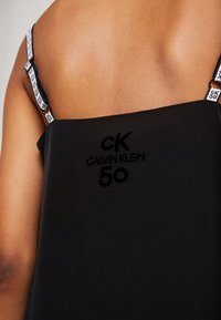Rückansicht einer Person, die ein schwarzes Calvin Klein Kleid mit markierten "ck 50" Trägern und Logo im oberen Rückenbereich trägt.