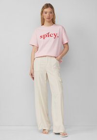 Helles rosa Baumwoll-T-Shirt mit dem roten Text "spicy." Kombiniert mit off-white weiter geschnittenen Cargo-Hosen, die über Taschen und eine strukturierte Oberfläche verfügen.
