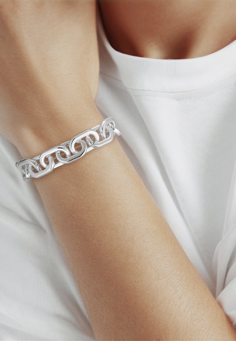 Close-up van een onderarm met een dikke zilveren schakelarmband, tegen een witte T-shirt met korte mouwen.