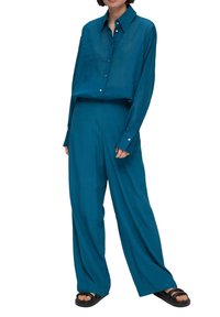 Turkos oversized skjorta med knappar, kombinerad med vida byxor i matchande färg. Mjukt tyg med en slät yta. Svarta sandaler kompletterar looken.