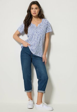 Femme debout, une main dans la poche, portant un chemisier blanc à fleurs à manches courtes, un jean bleu retroussé et des baskets blanches, devant un fond uni.