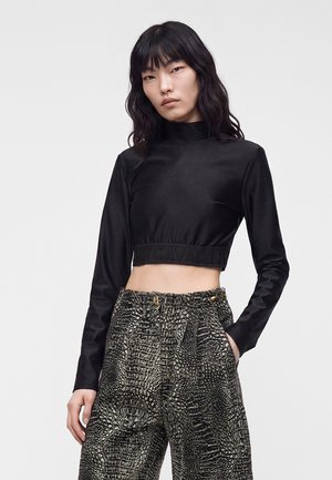 Jeune femme aux cheveux noirs ondulés portant un crop top noir à manches longues et un pantalon texturé métallique à taille haute, les mains dans les poches.