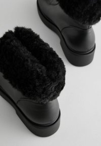Black boots featuring a plush, fuzzy black collar, smooth leather body, rounded toe, and thick rubber sole for added comfort.