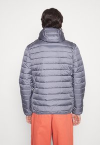 Personen står med ryggen til iført en grå polstret jakke med hætte og orange bukser mod en ensfarvet hvid baggrund.