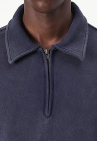 Maglioncino blu navy con colletto con zip, realizzato in tessuto morbido e strutturato. Presenta un orlo a coste e una vestibilità aderente intorno al collo.