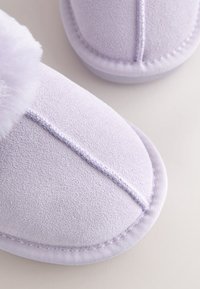 Gros plan sur des chaussons doux, violet clair, avec un tissu texturé et une finition duveteuse sur une surface claire.