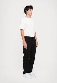 Zwarte broek met een gladde textuur, gecombineerd met een witte katoenen t-shirt en witte sneakers. Model staat in profiel tegen een effen achtergrond.
