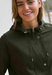 Giacca con cappuccio verde scuro, dotata di chiusura con zip, accessori in argento e cordoncino. Presenta una superficie liscia e una tasca frontale.